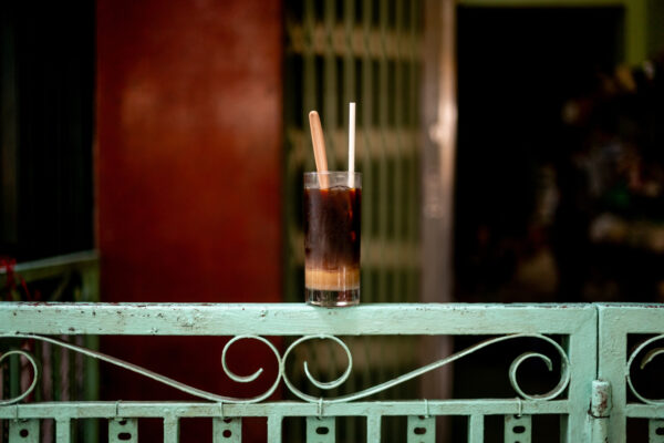 Tall glass of iced coffee with two straws resting on a weathered turquoise railing at dusk/place of a cafe.