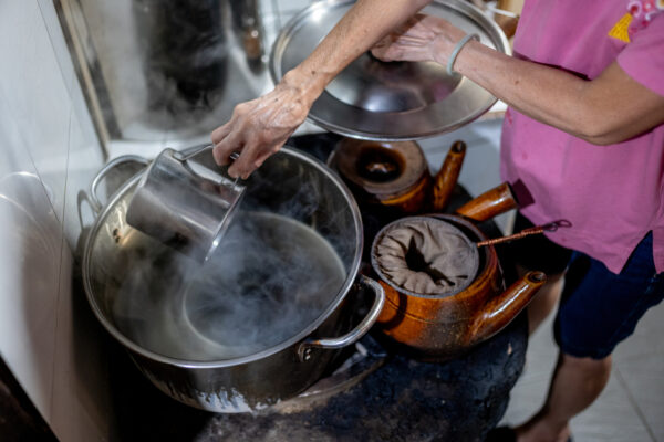 Person in a pink shirt pours steaming liquid from a metal pitcher into a large pot on a stove, kitchen steam rising from the pot.