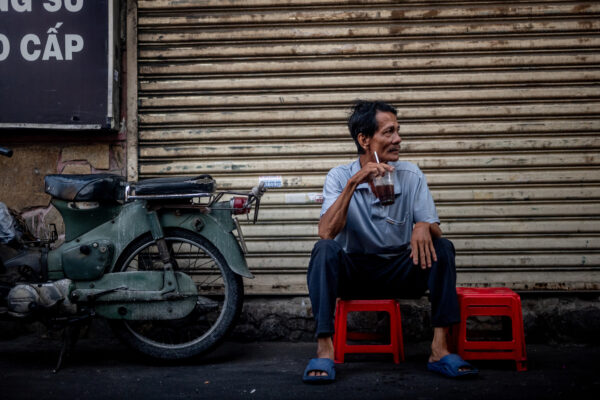 A man sits on red plastic stools in front of a closed metal shutter, sipping from a cup with a straw beside a dusty motorcycle.