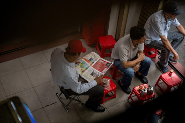 Three men sit on red plastic stools outside a shop; one reads a colorful magazine while others chat nearby, with drinks on a small red table.
