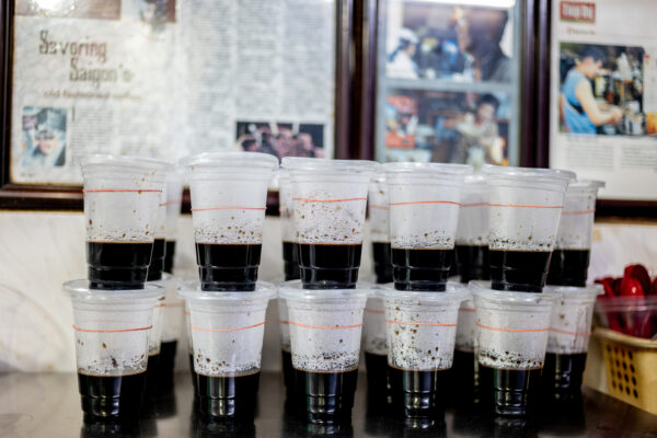 Rows of clear plastic cups filled with dark liquid on a table, stacked in two levels, with a red ring around each cup; blurred newspaper frames in the background.