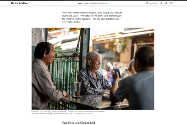 Three elderly men sit at a glass-topped street-side café table; one man concentrates on a smartphone while others chat in a busy urban setting.