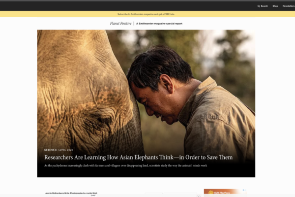 Man with a khaki shirt rests his head against the side of an Asian elephant in a natural setting, showing a close moment of study or bonding as part of field research.