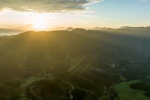 Aerial view of rolling hills at sunrise with warm sun rays over a valley and winding roads.