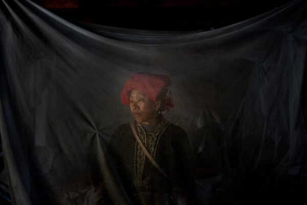 Portrait of a woman wearing a red headwrap and traditional embroidered clothing, seen through a dark sheer fabric veil.