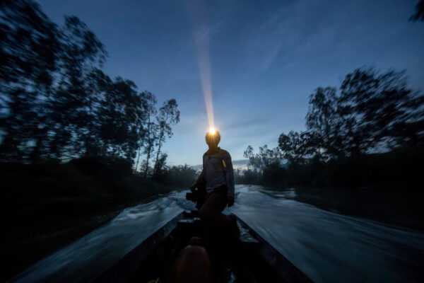 Person kneeling at the bow of a small boat on a river at dusk, wearing a headlamp that shines forward through the trees.