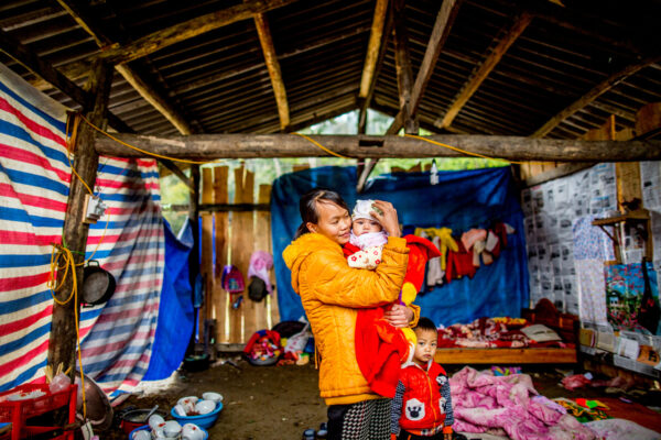 Mother in a yellow jacket cuddles a baby wrapped in red while a young boy stands nearby inside a makeshift shelter with colorful tarps and blankets nearby for warmth and shelter.