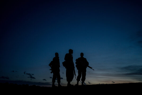 Silhouetted trio of soldiers with rifles standing on a hill at dusk against a blue gradient sky, horizon visible on the horizon edge.
