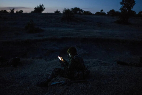 Soldier in camouflage gear sits on the ground at dusk, looking at a smartphone glowing in hand.