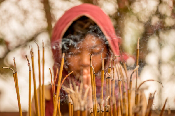 Incense sticks burn in the foreground while a person in a red hooded garment sits blurred in the background, with wisps of smoke rising.