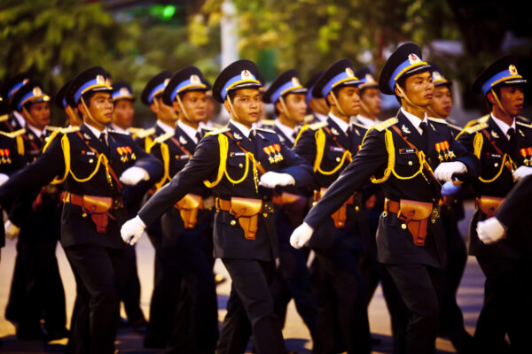 Group of soldiers in ceremonial dress uniforms marching in formation during a parade, wearing dark blue outfits with gold cords and medals.