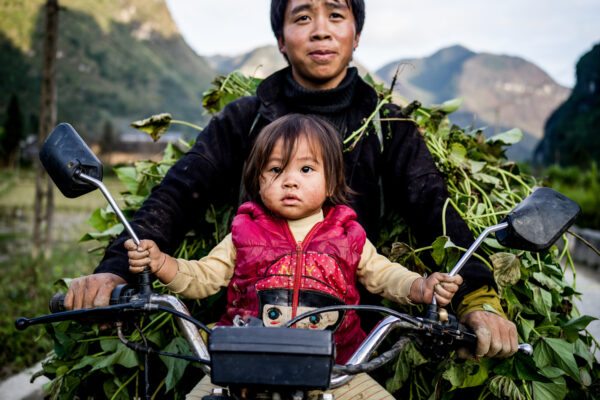 Man and child ride a motorbike piled with leafy branches, in a rural valley setting.