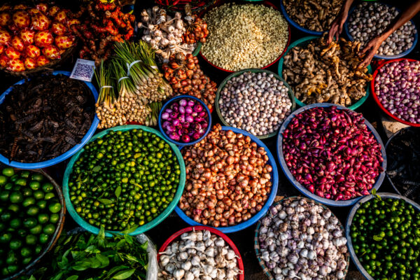 Colorful market stall with bowls of limes, onions, shallots, garlic, ginger, lemongrass, and assorted peppers and roots arranged in vibrant baskets
