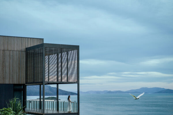 Woman in white dress standing on a balcony of a modern building overlooking a calm sea and distant mountains in the background.
