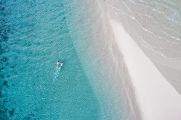 Aerial view of turquoise sea meeting a white-sand beach; two people on paddleboards near the shore.