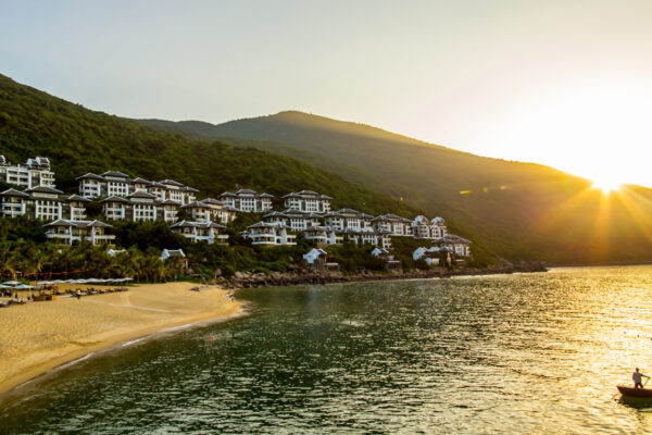 Resort buildings on a green hillside overlook a sandy beach and calm water at sunset, with a small boat in the foreground.