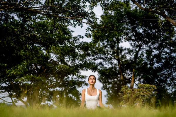 Person meditating outdoors in a grassy field beneath leafy trees, eyes closed, wearing a white tank top.