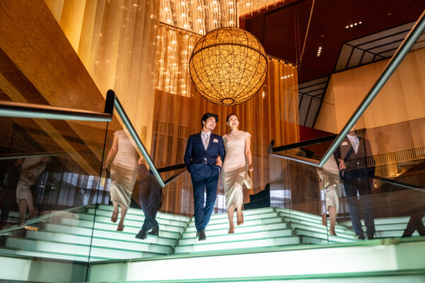 Pair of well-dressed adults descending glass stairs in a modern lobby with a large hanging orb chandelier.