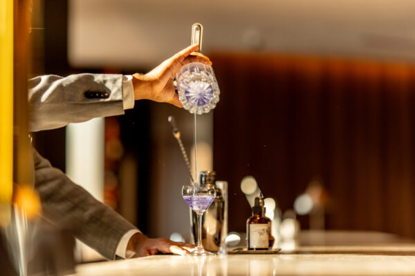 Bartender in a suit pours clear liquid from a glass bottle into a stemmed coupe glass at a bar.