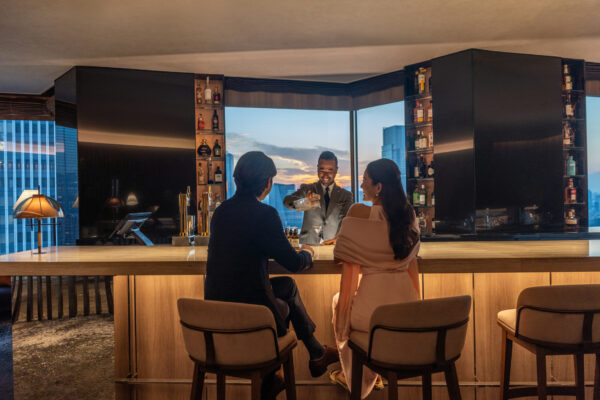 Bartender in a suit pours a drink for a couple at a high wooden bar with city skyline visible through large windows behind them.