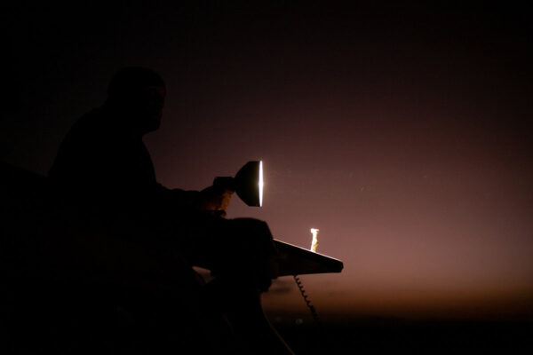 Silhouette of a person on a dark vehicle at night, holding a bright handheld spotlight pointing forward.