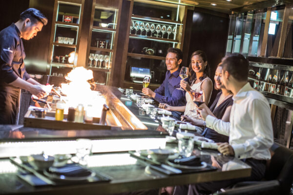 Chef grilling on a teppanyaki grill as flames rise, while five diners at the counter toast with wine and smiles.
