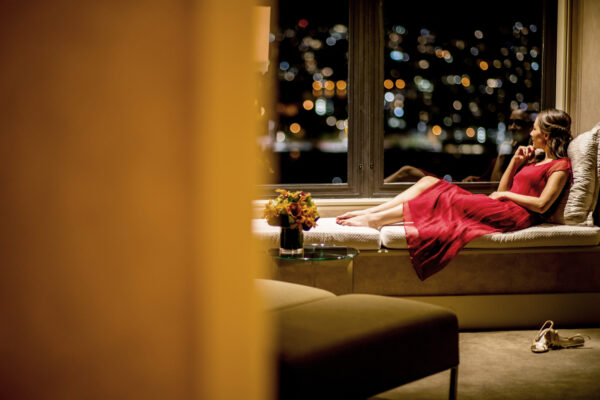 Woman in a red dress relaxing on a window seat, gazing at blurred city lights at night, with a vase of flowers nearby.