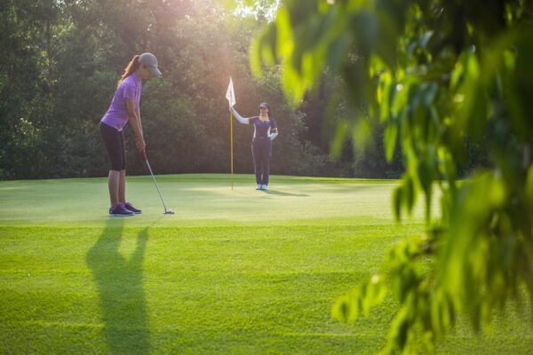 Two women on a sunny golf green: one putstling while the other stands by the hole with the flag nearby, surrounded by trees.
