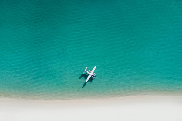 A white seaplane floating on calm turquoise water just off a pale sandy beach, aerial view.