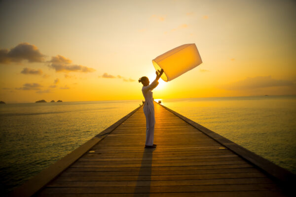 Person on a wooden pier at sunset releasing a large yellow lantern into the air over calm water.