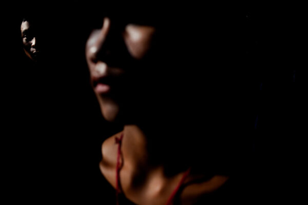 Dark, low-light portrait with a partially visible face peering from the left; a blurred figure with red straps dominates the foreground.