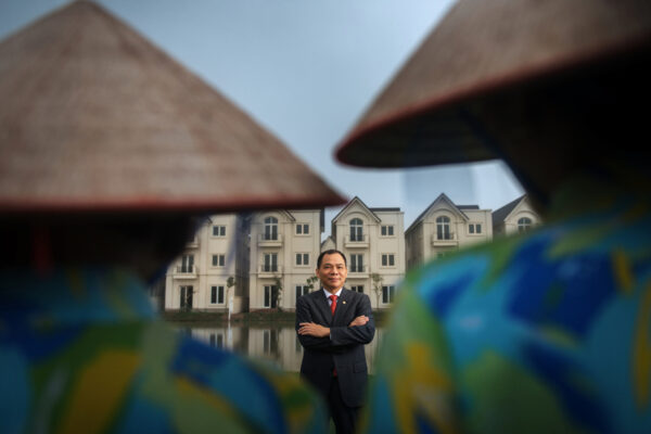 Man in a suit with arms crossed standing in front of a row of beige apartment buildings, framed by blurred foreground elements.