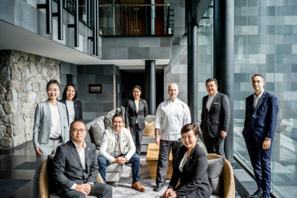 Group of nine professionals in business attire posing in a modern glass-walled lobby.