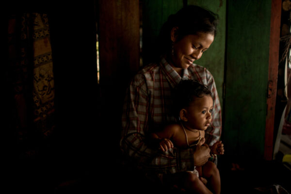 Mother in a plaid shirt cradling her baby in a dimly lit room, warm light on their faces.”