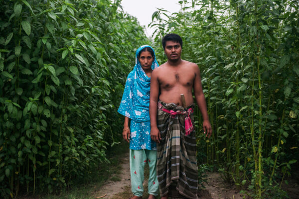 A shirtless man in a plaid lungi stands on a dirt path between tall green plants, with a woman in a blue patterned headscarf beside him.