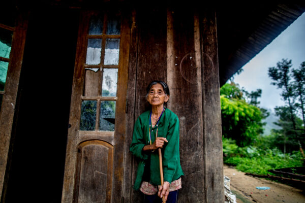 Old woman in a green jacket leaning on a wooden walking stick outside a weathered door. It conveys a rural, elderly figure at a rustic home.