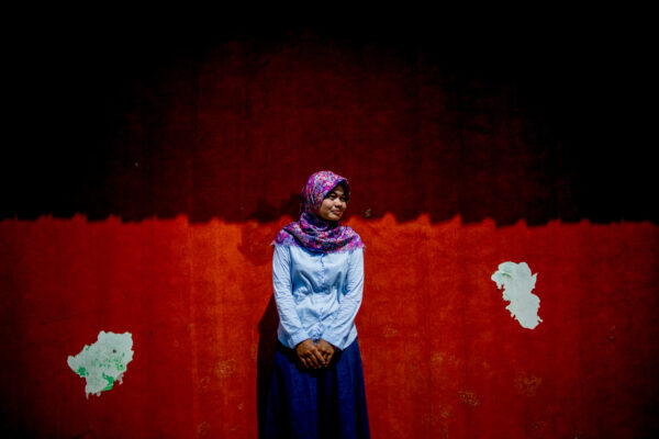 Woman in a light blue blouse and colorful hijab stands centered against a two-tone red wall with peeling white patches in the foreground border area.