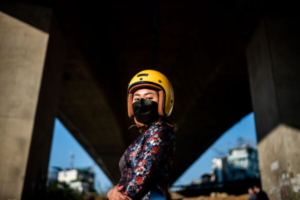 Person in a yellow helmet and floral long-sleeve shirt wears a black face mask under a concrete overpass, looking at the camera.