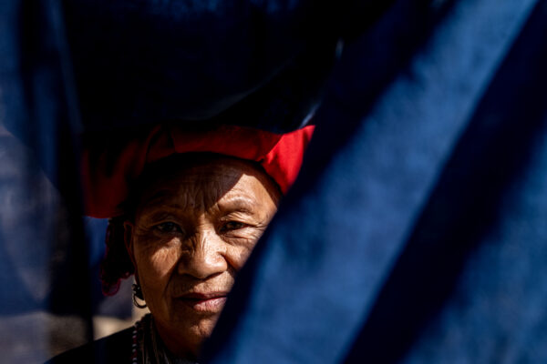 Older woman wearing a red headscarf peeks through dark blue fabric, with a calm, intimate gaze.