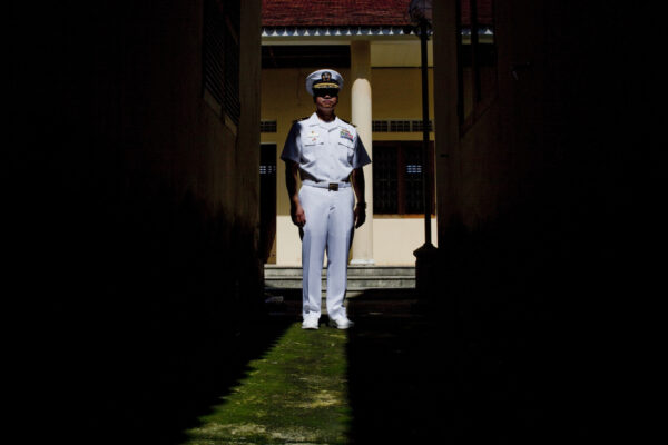 A military officer in a white dress uniform stands in a sunlit doorway between dark walls.