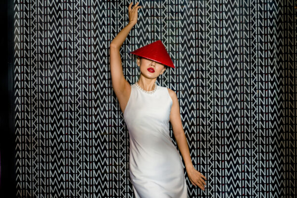 Model in a white satin dress with a red cone hat and pearl necklace, posing with one arm raised against a geometric patterned backdrop.