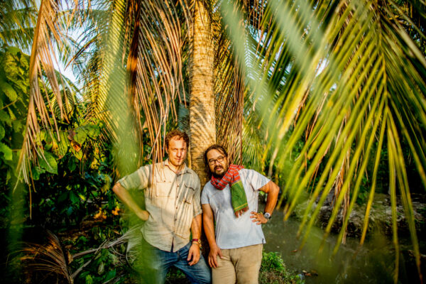 Two men pose leaning against a palm tree in a sunny tropical garden. A beige shirt and a light gray shirt with a scarf, both relaxed.