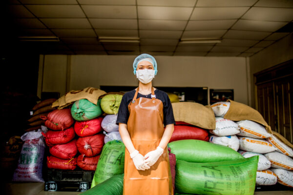 Worker wearing a blue hairnet, white mask, and orange apron stands with hands clasped in front of stacked colorful bags in a warehouse