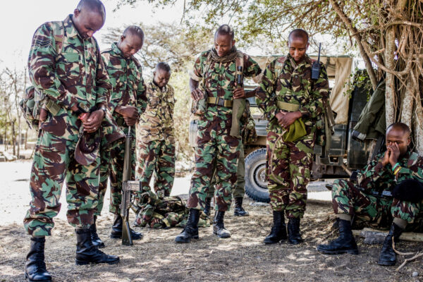 Group of soldiers in camouflage uniforms standing with rifles, heads bowed in a dusty outdoor area near a vehicle and trees, military gear visible.