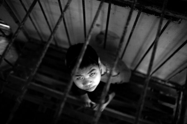 Black-and-white image of a young boy looking up at the camera from a dim stairwell, framed by rusted metal railings.