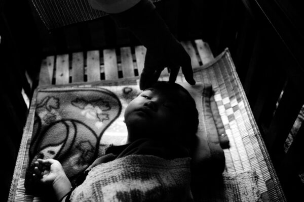 Infant asleep in a crib as a caregiver gently rests a hand on the baby's head.