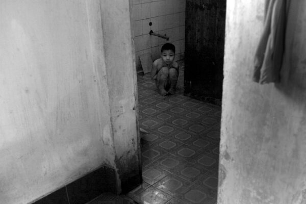 Child crouching on the bathroom floor in a corner, looking toward the camera.