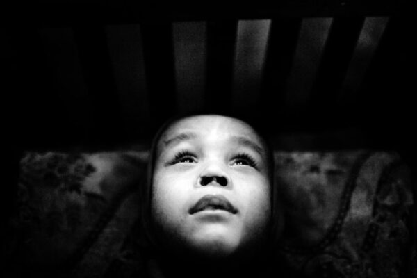 Black-and-white photo of a baby lying on its back, gazing upward with wide eyes; crib slats are visible above.
