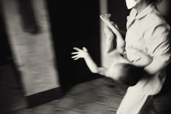 Blurred black-and-white scene of a masked person holding a child, with a reaching hand toward the left amid motion blur