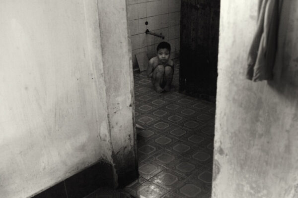 Child crouches on a tiled bathroom floor in a doorway, holding knees and looking toward the camera in a dim, worn room with a faucet on the wall behind.